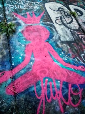Graffiti image of an all pink silhouette of a person sitting cross legged w/ their hands outstretched as if in meditation. They have a crown above their head.